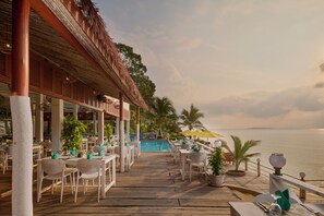 A wooden deck with tables and chairs, a pool, and a view of the sea.