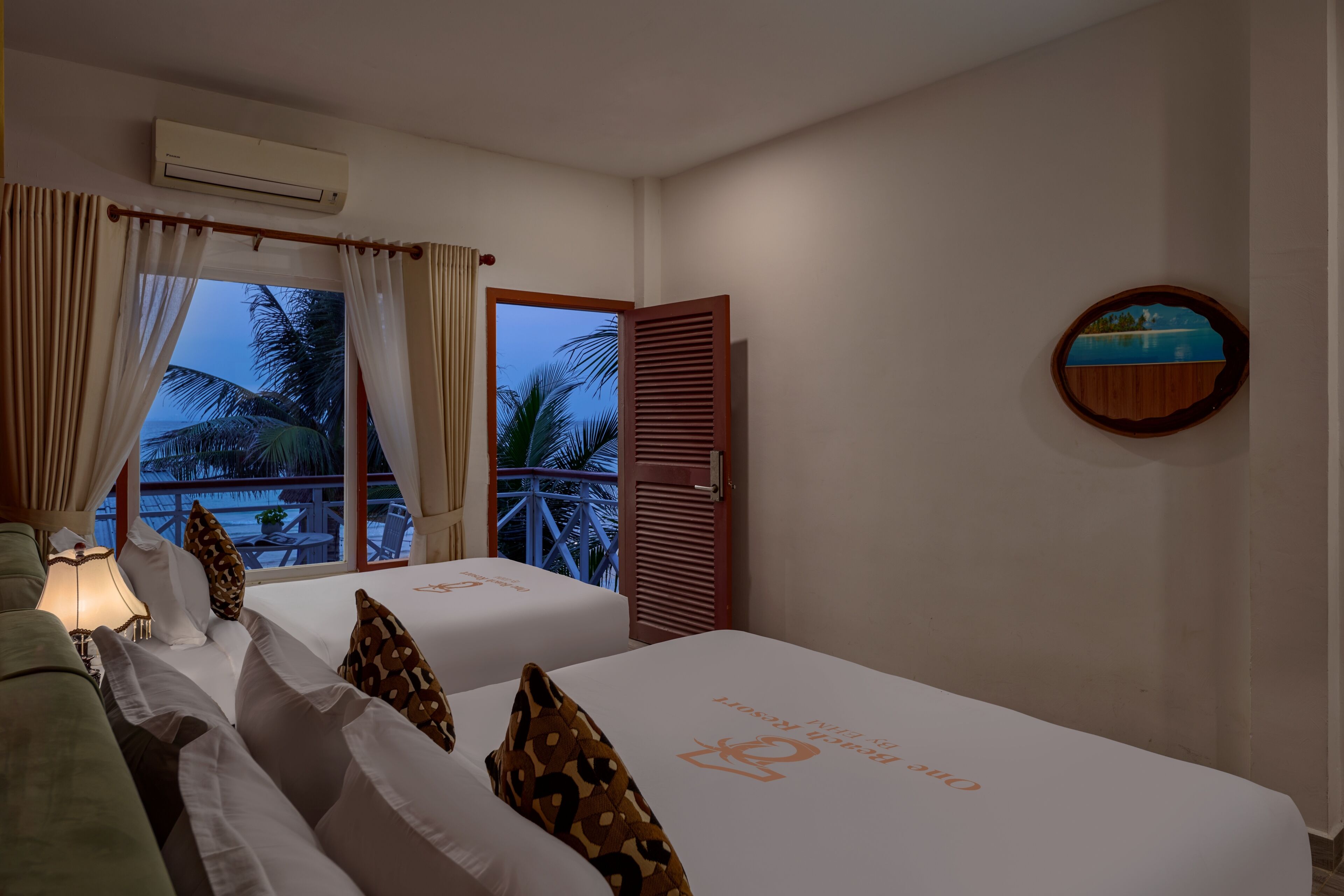 A hotel room with two beds, a balcony with a view of palm trees, an air conditioning unit, and a wooden-framed picture on the wall.