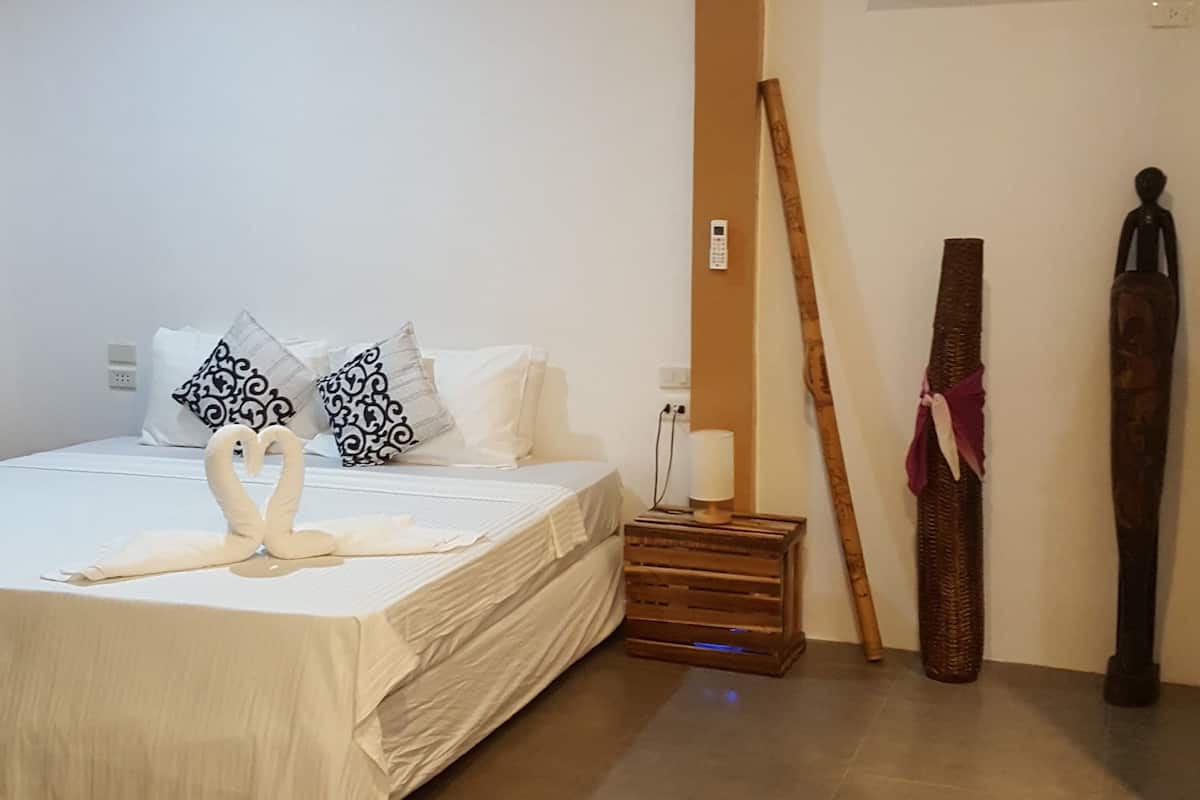A bedroom with a bed, pillows, a bedside table, a lamp, a wall-mounted air conditioner, and a wooden object leaning against the wall.