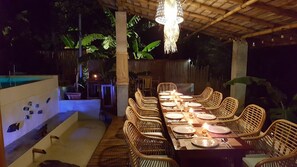 An outdoor dining area with a long table set for six, surrounded by wicker chairs, under a wooden pergola with a hanging light fixture.