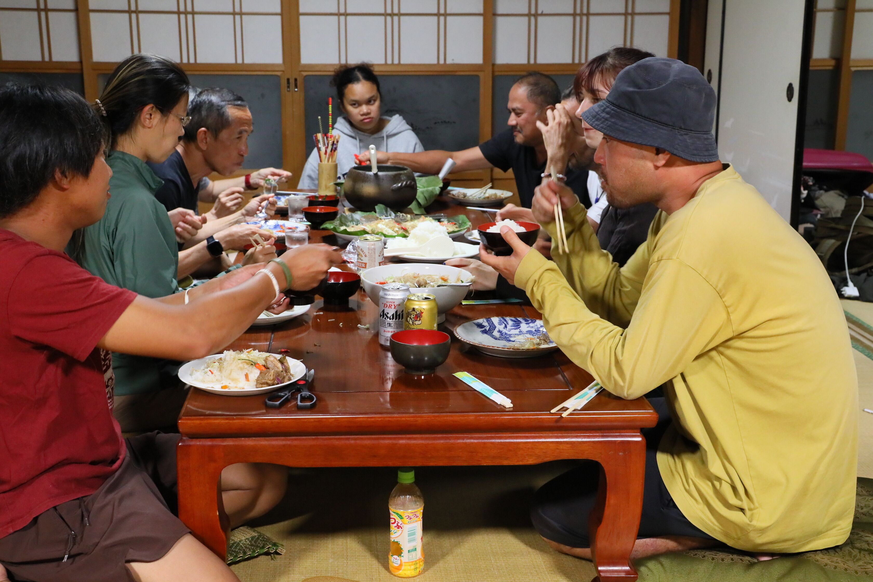 Um grupo de pessoas sentadas em volta de uma mesa de madeira, com vários pratos e uma garrafa de refrigerante.