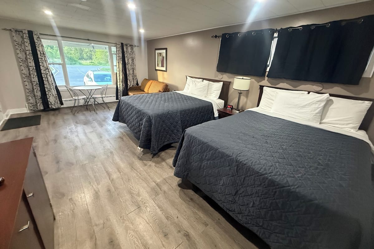 Habitación de hotel con dos camas, un escritorio, una silla, un ventanal con cortinas, una lámpara y un cuadro en la pared.