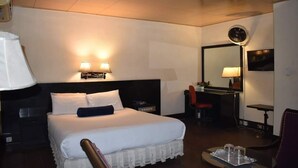 Habitación de hotel con una cama grande, lámparas de noche, un escritorio con silla, un espejo y una pequeña mesa con vasos.