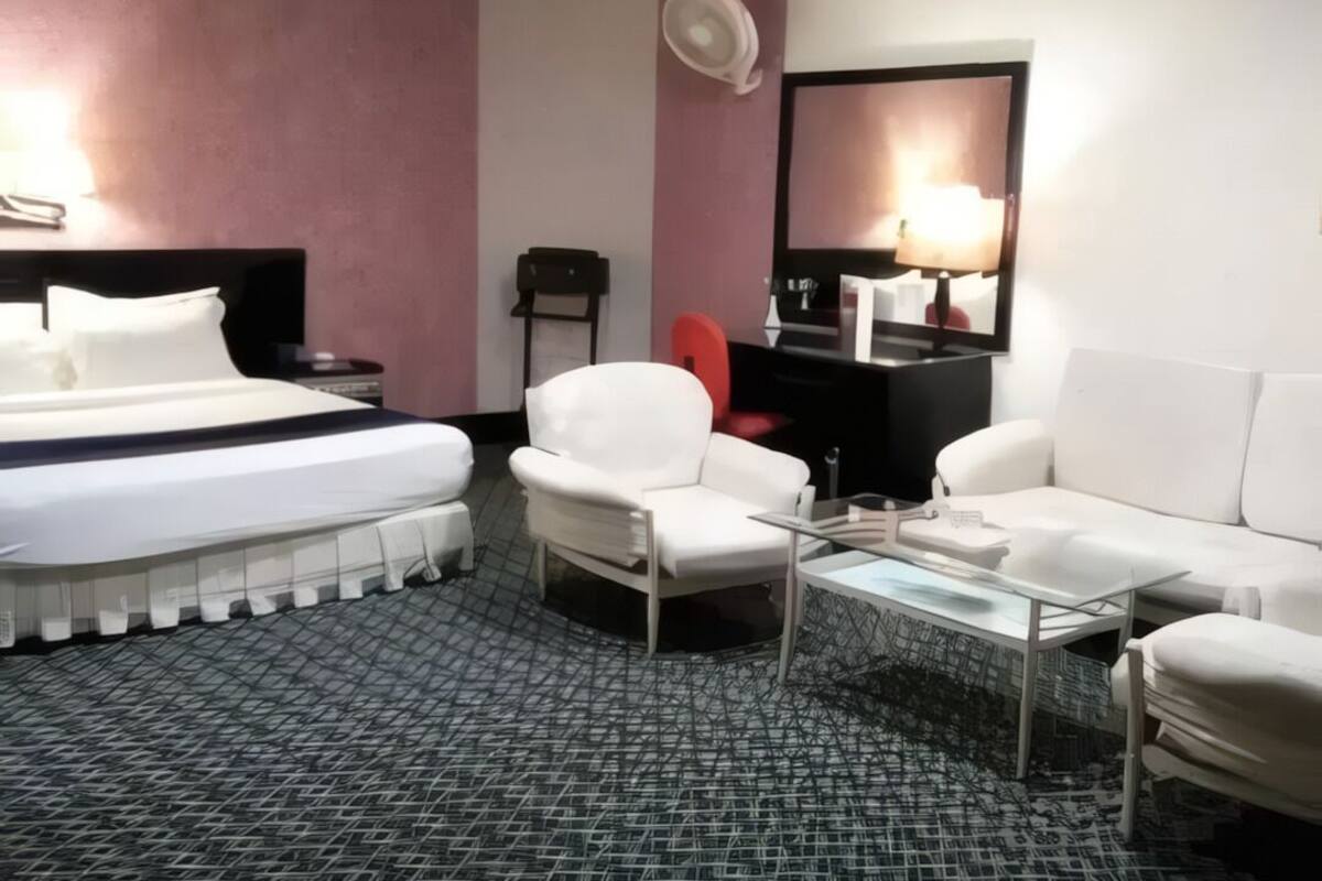A hotel room with a bed, two white armchairs, a glass coffee table, a red chair, a black desk, and a mirror on the wall.