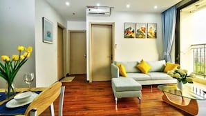 Een moderne woonkamer met een bank, een voetenbankje en een glazen salontafel. Er is een eethoek met een houten tafel en stoelen. De kamer heeft houten vloeren en een groot raam met gordijnen.
