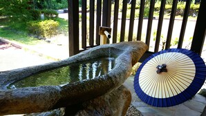 A stone water feature with a wooden spout, a traditional umbrella, and a wooden fence in the background. - Gensen to Hanare no Oyado Tsuki (Ito)