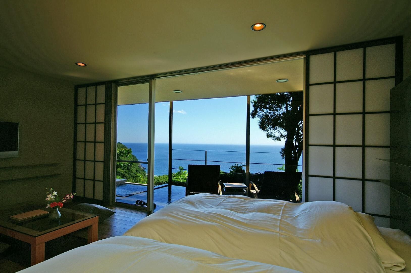 Een slaapkamer met een groot bed, uitzicht op de oceaan en een schuifdeur naar een balkon met stoelen.