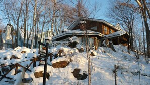 Uma cabana de madeira com telhado coberto de neve, rodeada por árvores e pedras.