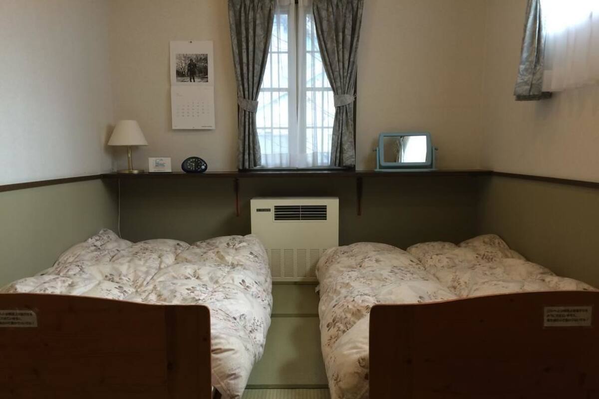 Ein Zimmer mit zwei Einzelbetten, einem Fenster mit Vorhängen, einem wandmontierten Kalender, einem kleinen Tisch mit Lampe sowie einem an der Wand befestigten Fernseher.