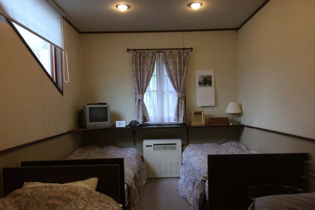 Ein kleiner, gemütlicher Raum mit zwei Einzelbetten, einem Fernseher auf einem Regal, einem Fenster mit Vorhängen und einem Kalender an der Wand.