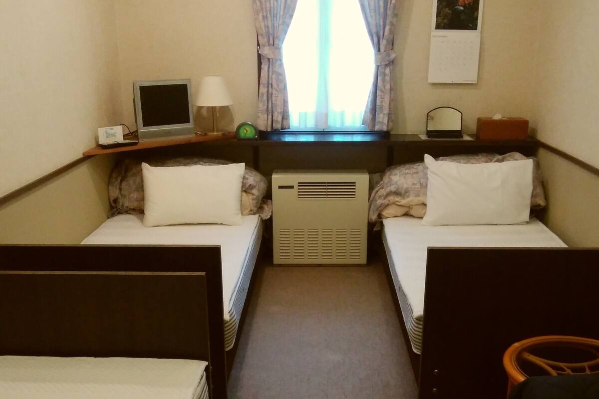 Ein Hotelzimmer mit zwei Einzelbetten, einem Schreibtisch mit Computer, einem kleinen Tisch mit Lampe, einem Fenster mit Vorhängen und einem Wandkalender.