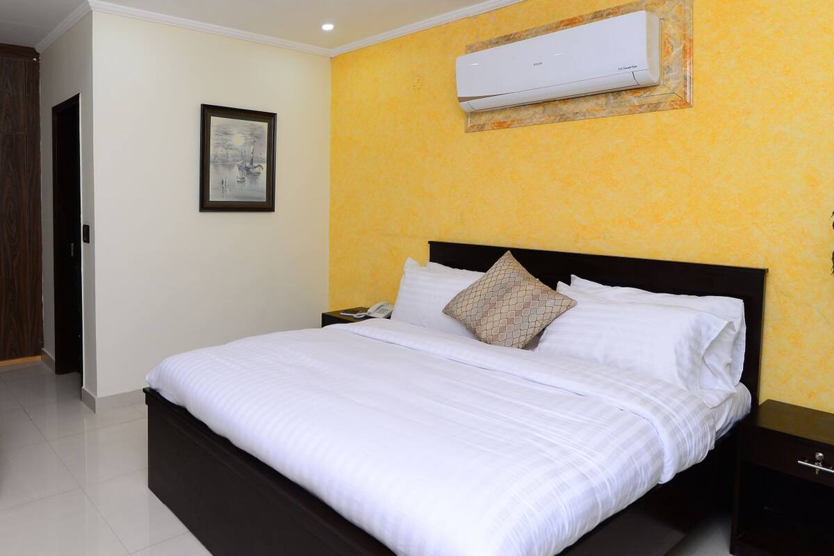 A hotel room with a bed, a wardrobe, a bedside table, and an air conditioning unit on the wall.