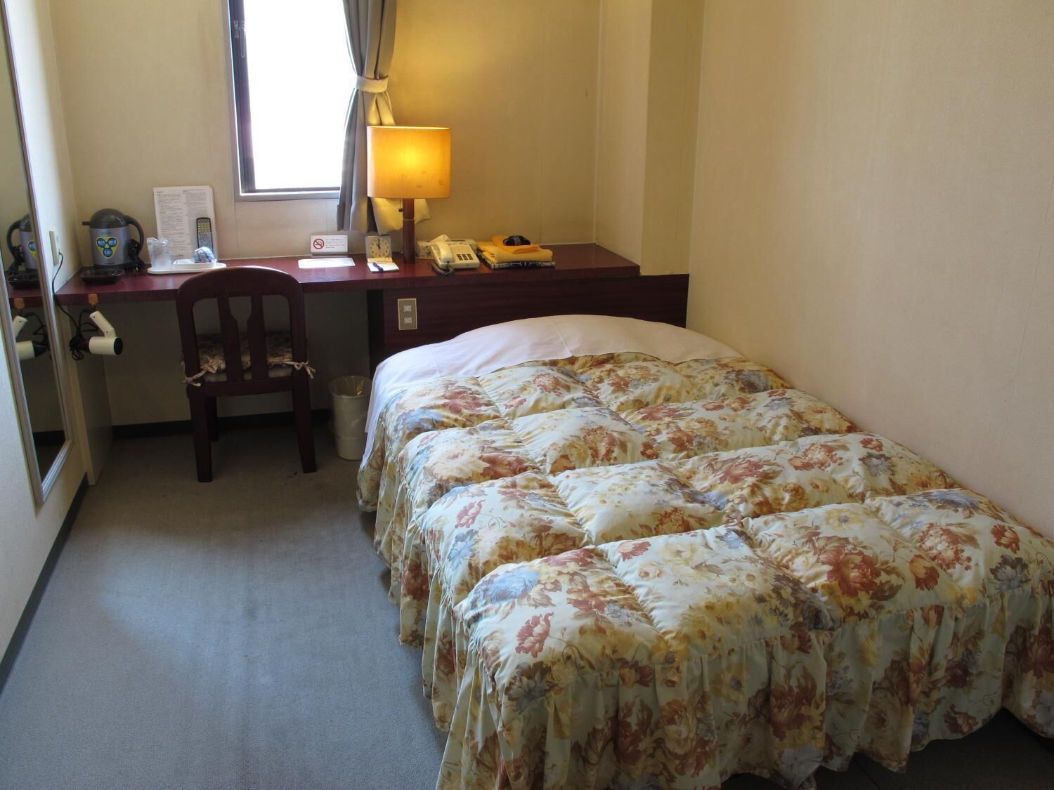 Et hotelværelse med en seng, et skrivebord med lampe, en stol og et vindue.