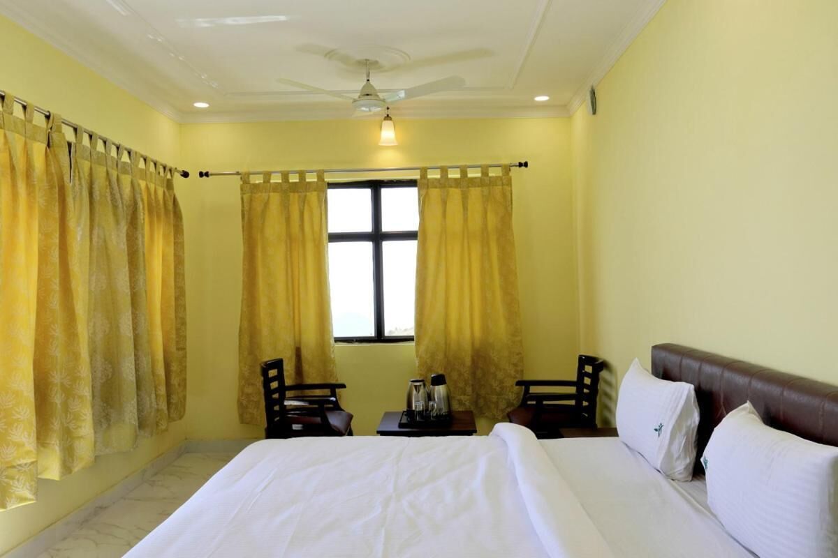 A hotel room with a bed, two chairs, a small table with a kettle and cups, and a window with curtains.