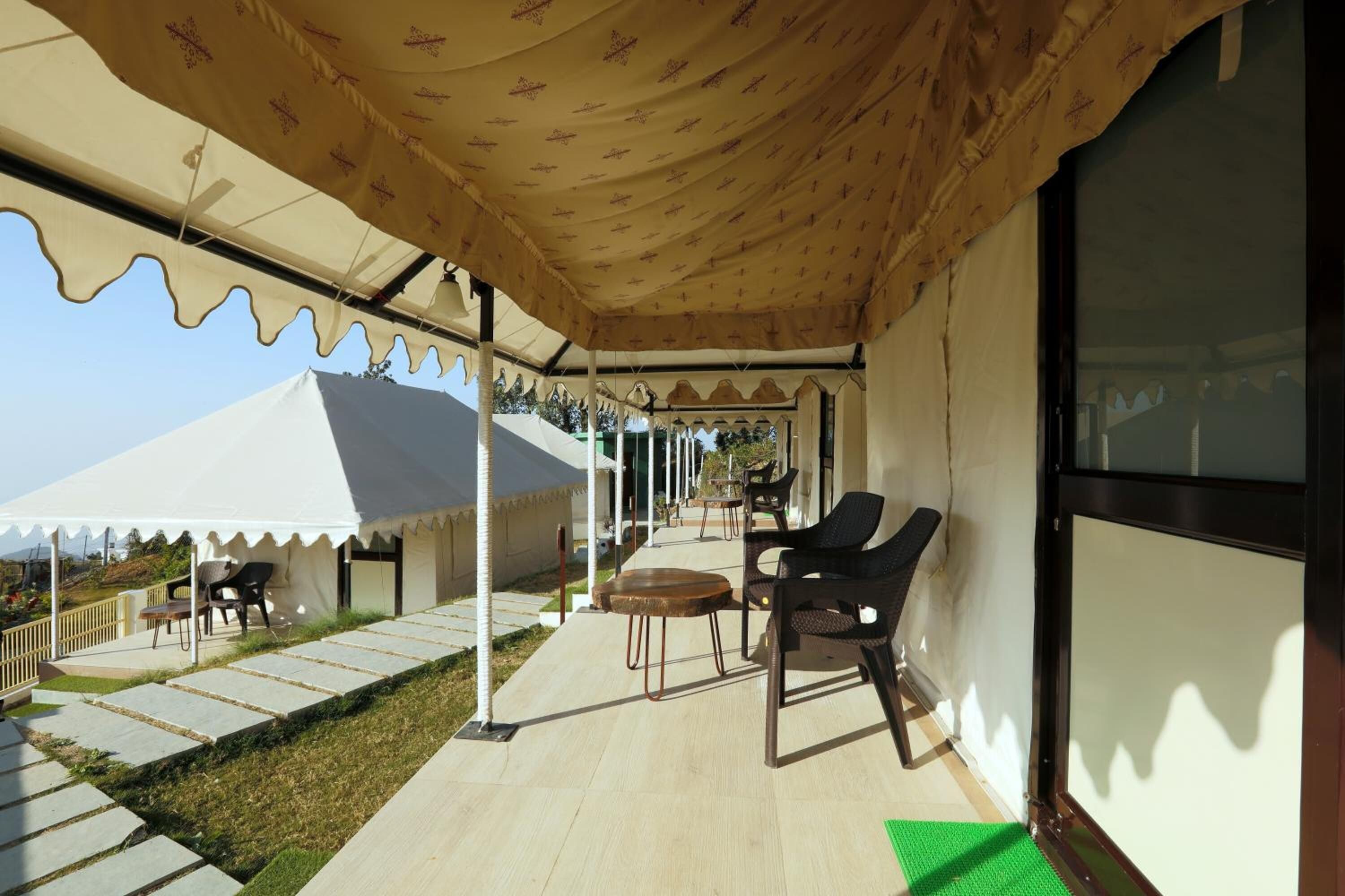 A covered outdoor seating area with tables and chairs, a tent in the background, and a clear sky.