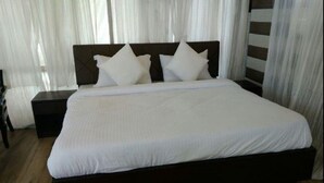 A neatly made bed with a dark headboard and white bedding, a nightstand to the left, and sheer curtains in the background. - Charekh Food & Forest Resort (Lansdowne)