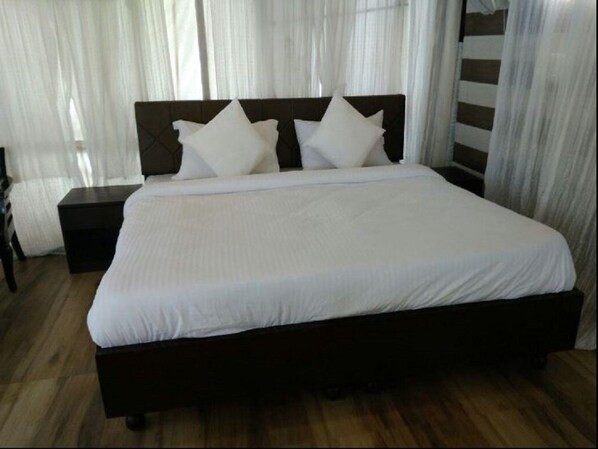 A neatly made bed with a dark headboard and white bedding, a nightstand to the left, and sheer curtains in the background.