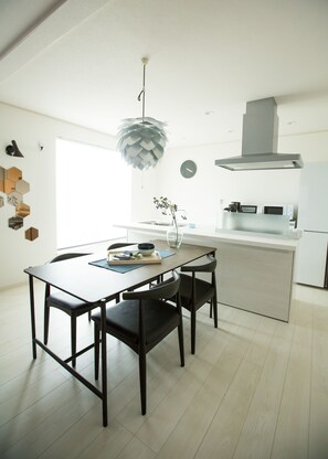 A modern kitchen with a dining area, featuring a wooden table, dark chairs, a unique pendant light, and a minimalist design. - Jikukan Hare (Onna)