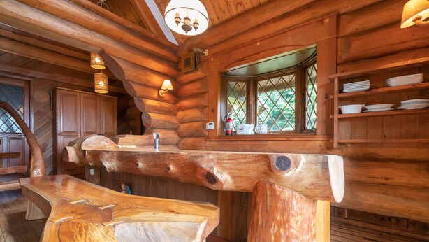 Ein rustikales Holzhäuschen im Inneren, mit einem großen Holztisch, Holzregalen und einem Fenster mit Blick auf grüne Landschaft.