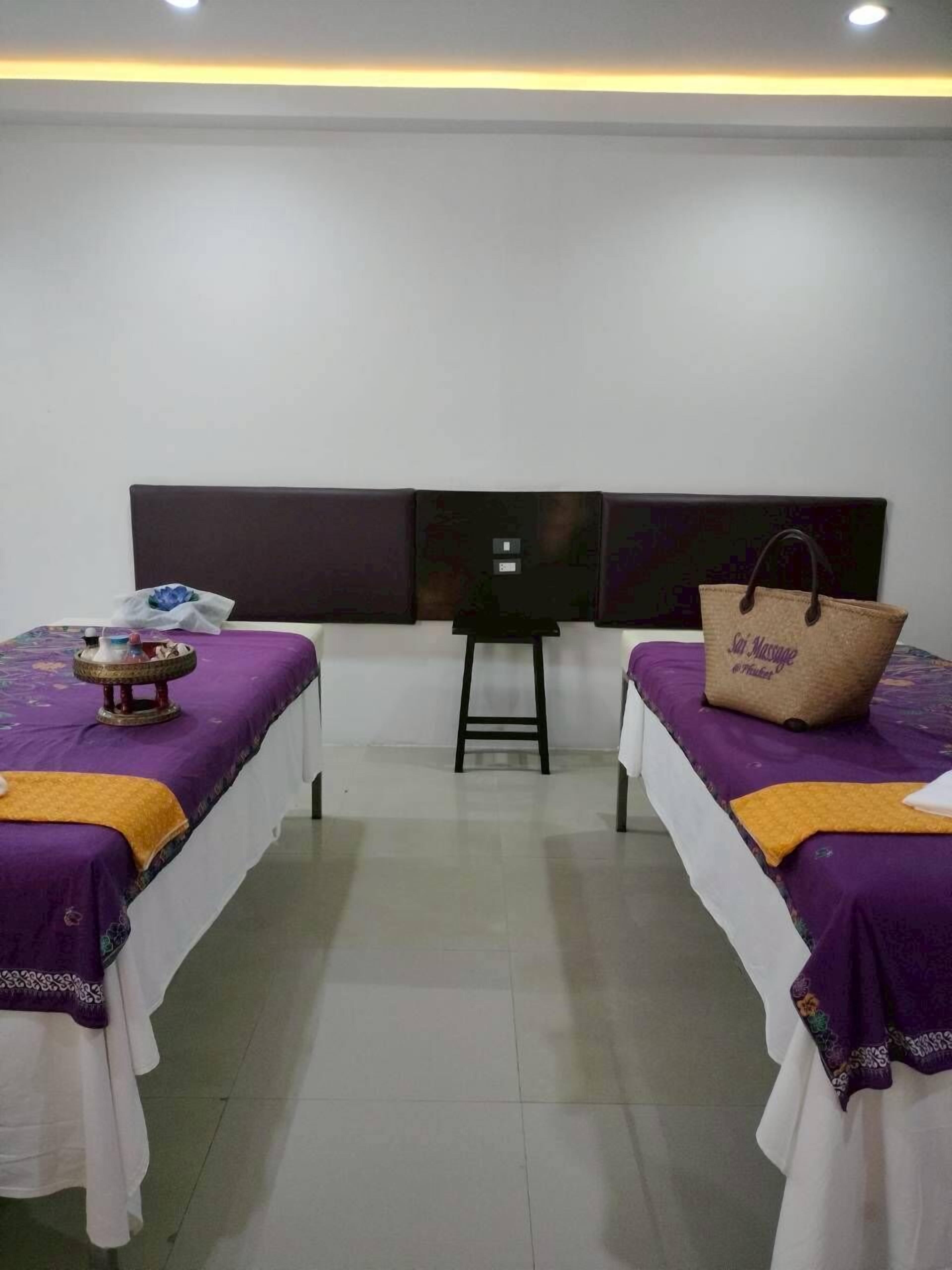 Habitación con dos camas, cada una con una colcha morada y una sábana blanca. Se observa una pequeña mesa con un objeto decorativo en la cama de la izquierda y un bolso marrón en la cama de la derecha.