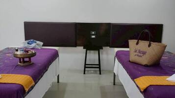 Habitación con dos camas, cada una con una colcha morada y una sábana blanca. Se observa una pequeña mesa con un objeto decorativo en la cama de la izquierda y un bolso marrón en la cama de la derecha.