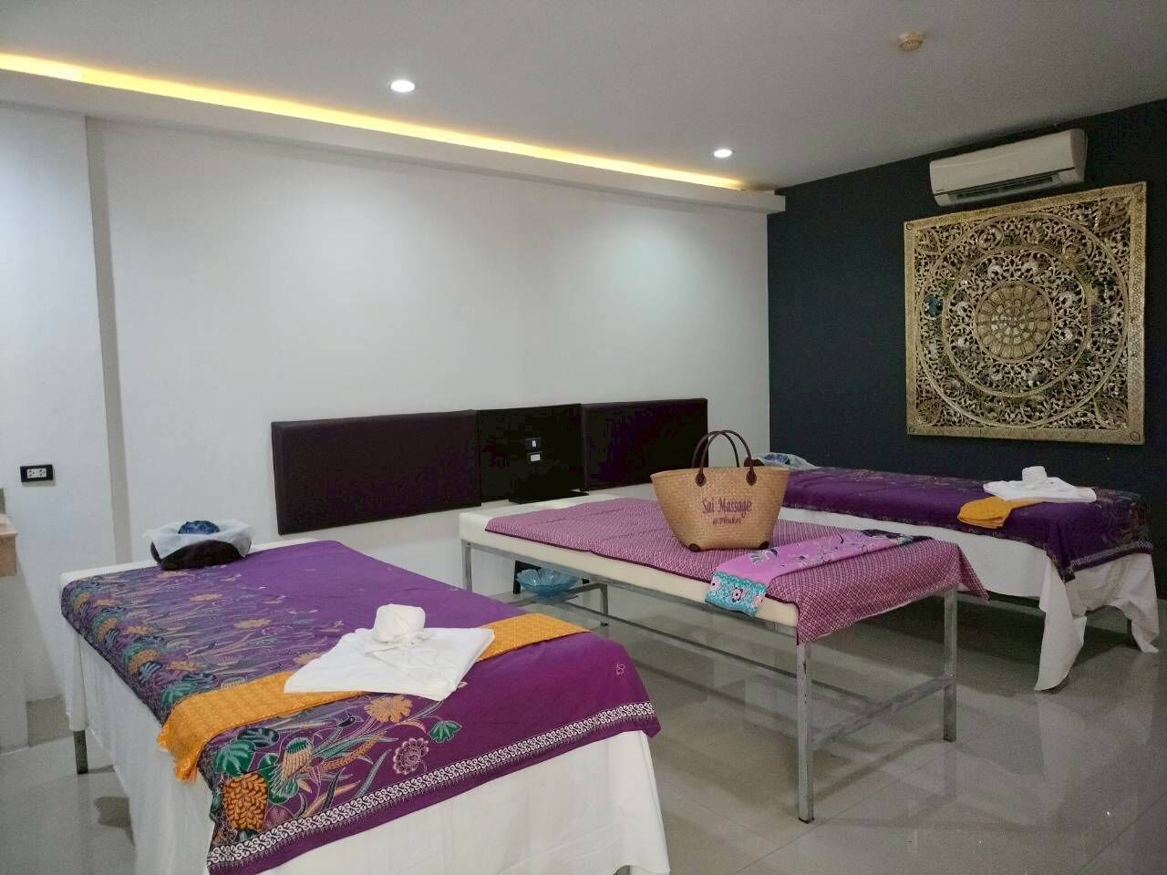 A room with two massage tables, each with a purple blanket and a white towel. There is a large decorative wall piece above the tables and a wall-mounted air conditioner.