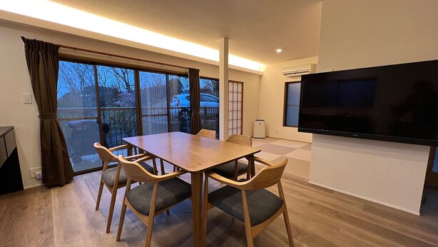 Una sala de estar moderna con una mesa y sillas de madera para comer, un televisor de pantalla plana grande y vistas a través de un ventanal.