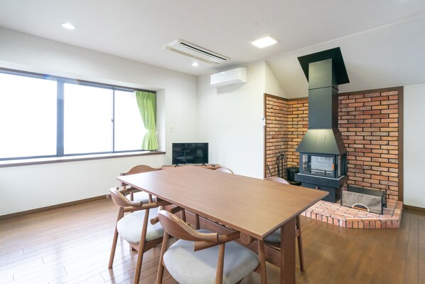 Una habitación con una mesa de comedor y sillas de madera, un televisor, una chimenea de ladrillo y una ventana con cortinas.