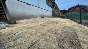Un patio trasero residencial con un muro de hormigón, una valla metálica y una casa de color azul.