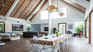 Une pièce spacieuse avec une longue table à manger en bois, entourée de chaises transparentes, un billard et une grande fenêtre laissant entrer la lumière naturelle.