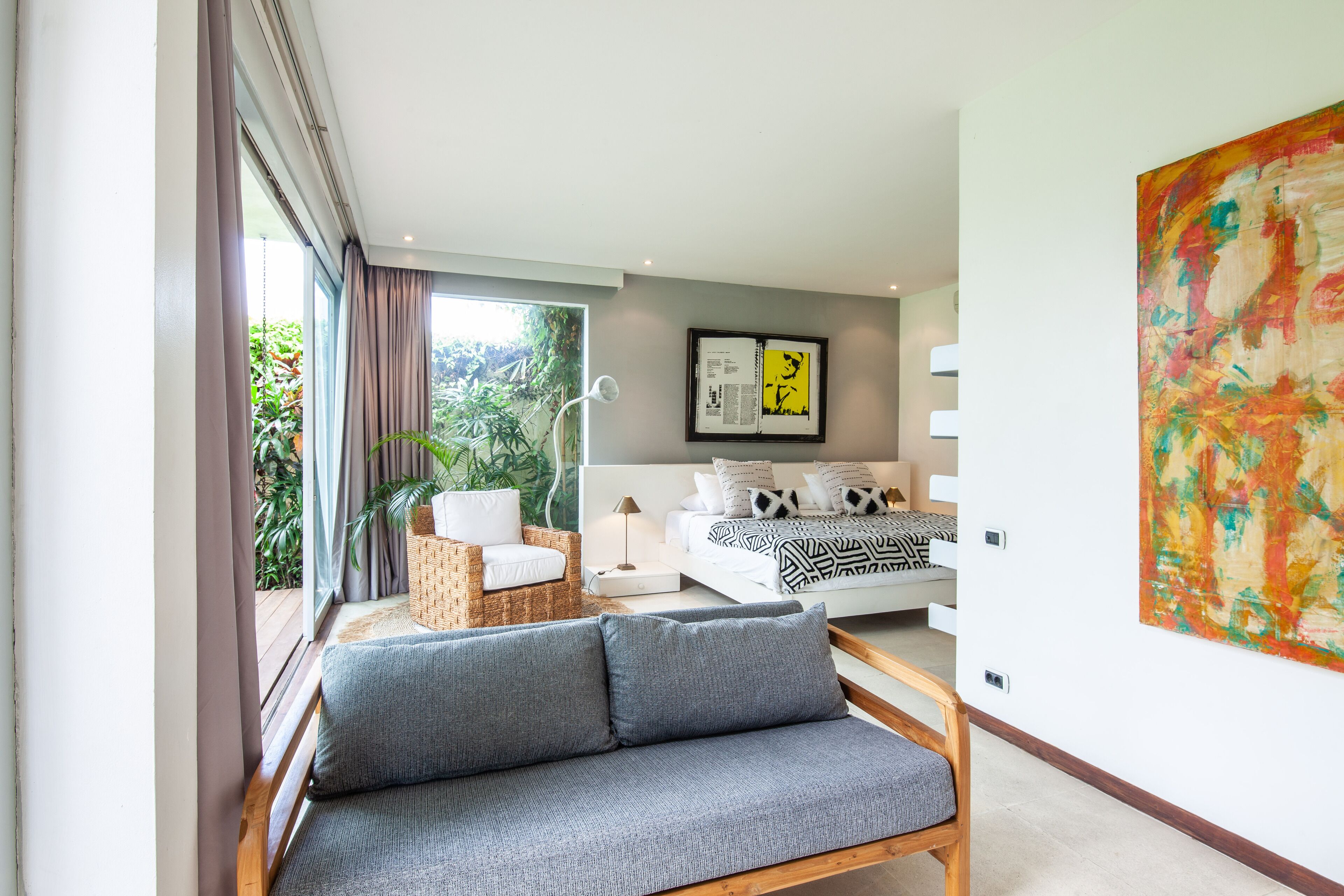 A modern bedroom with a large bed, a sofa, and a dining table. There is a window with curtains and a view of greenery outside.