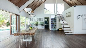 Une cuisine spacieuse avec une longue table en bois et des chaises transparentes, un escalier avec une rampe et une vue sur une piscine à l’extérieur.