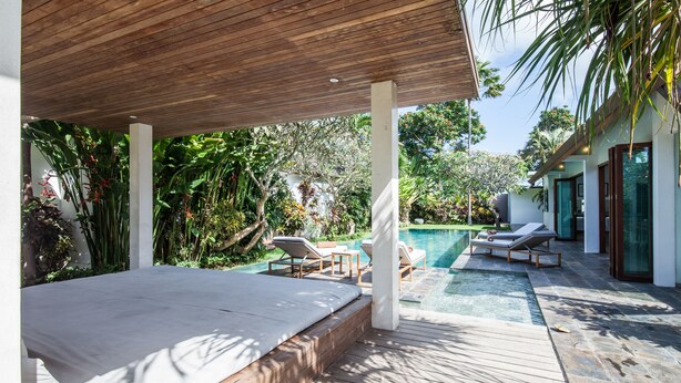 Une terrasse en bois avec un lit et des chaises longues, une piscine et une végétation luxuriante.