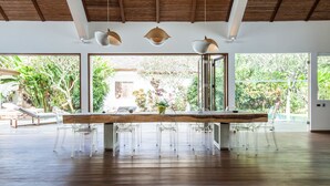 Une pièce spacieuse avec une longue table en bois, des chaises en plastique transparent et des luminaires suspendus. De grandes fenêtres offrent une vue sur la verdure environnante.
