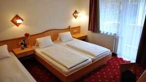 A hotel room with two beds, a wooden headboard, a nightstand with a lamp, a window with curtains, and a radiator. - Pension Posauner Dorfgastein (Dorfgastein)