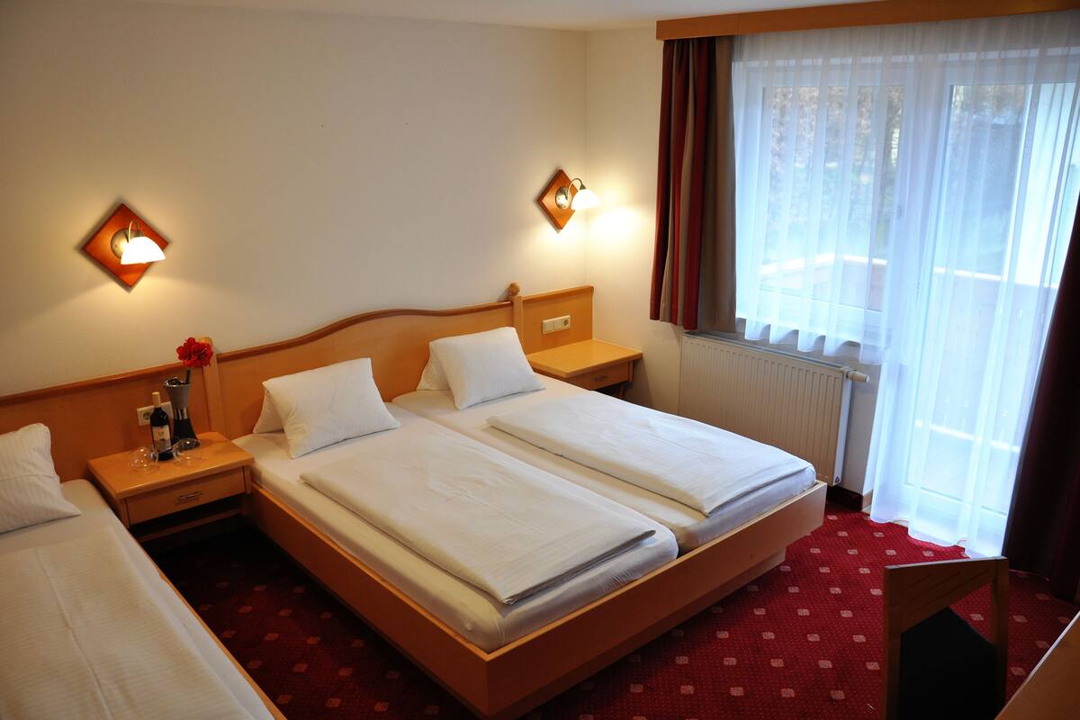 A hotel room with two beds, a wooden headboard, a nightstand with a lamp, a window with curtains, and a radiator.