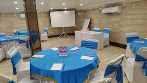 Una sala de conferencias con mesas redondas, sillas blancas con cintas azules, una pantalla de proyector y un equipo de aire acondicionado en la pared.