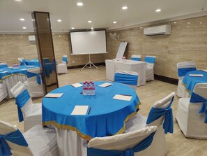 A conference room with round tables, white chairs with blue sashes, a projector screen, and an air conditioning unit. - Hotel Z Suites Near IGI Airport Delhi (New Delhi)