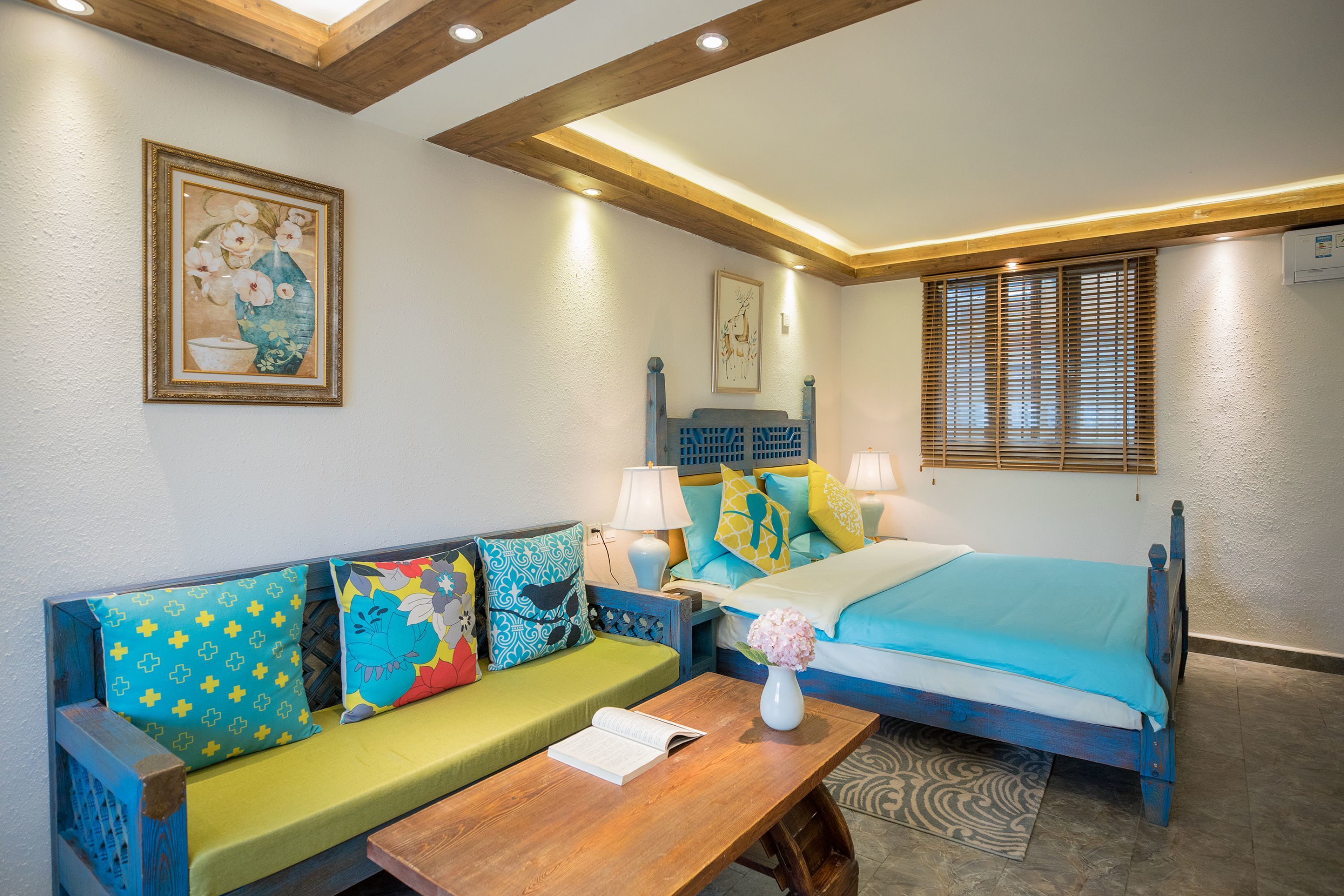 A bedroom with a bed, a wooden nightstand, a table with a book, a sofa with patterned cushions, and a wall-mounted picture.