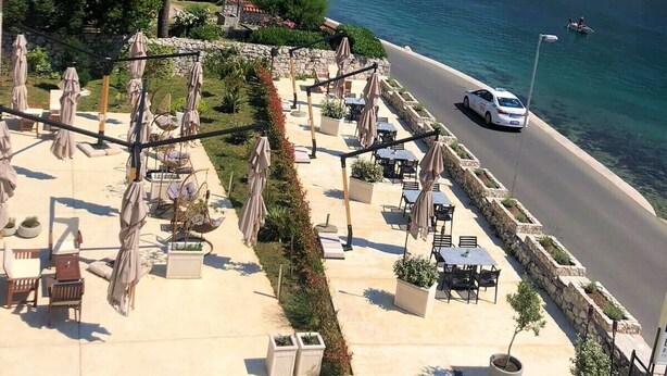 A coastal outdoor seating area with tables and umbrellas, overlooking a body of water and mountains.