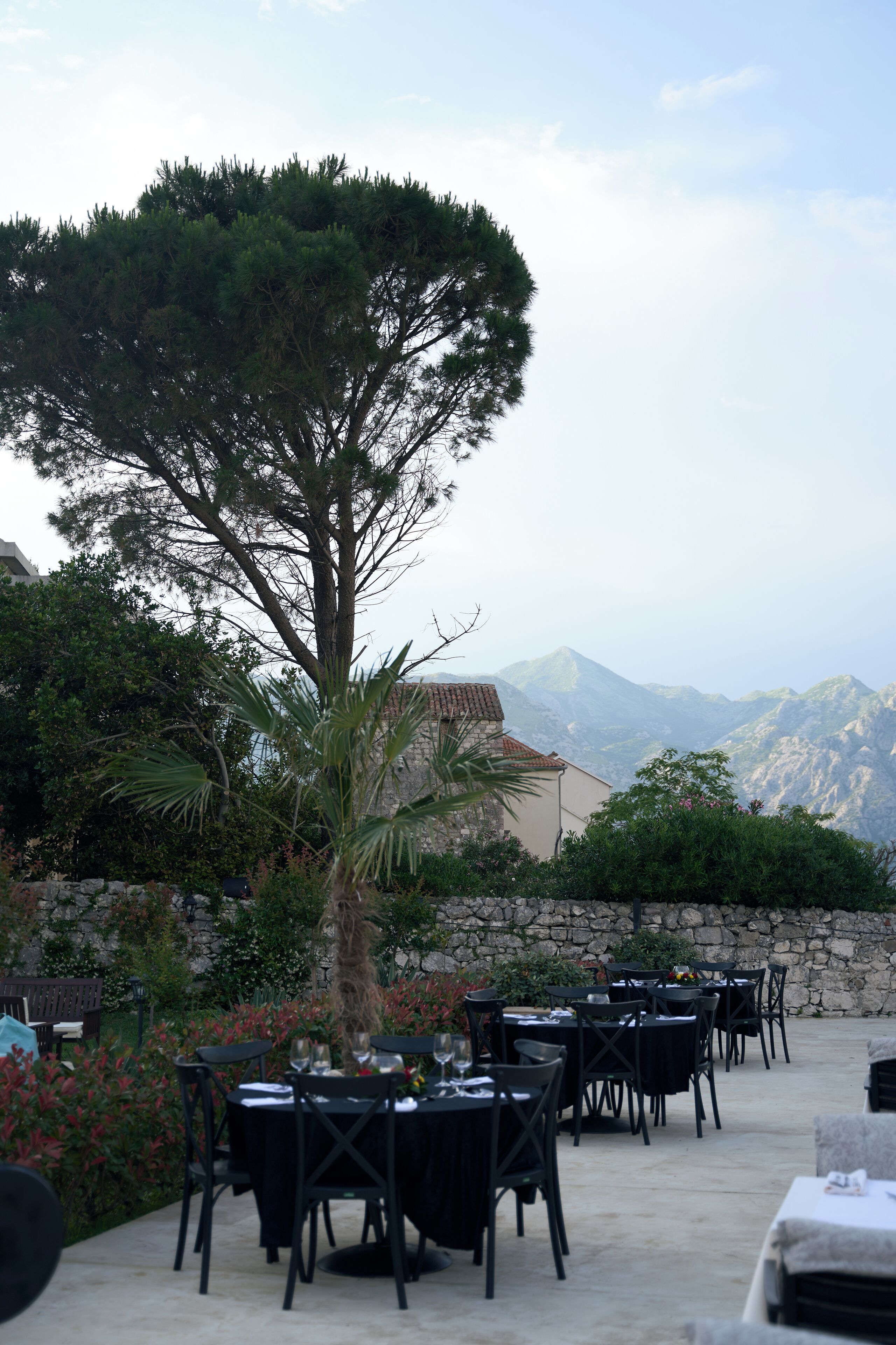 An outdoor dining area with tables and chairs, surrounded by greenery and a view of mountains.