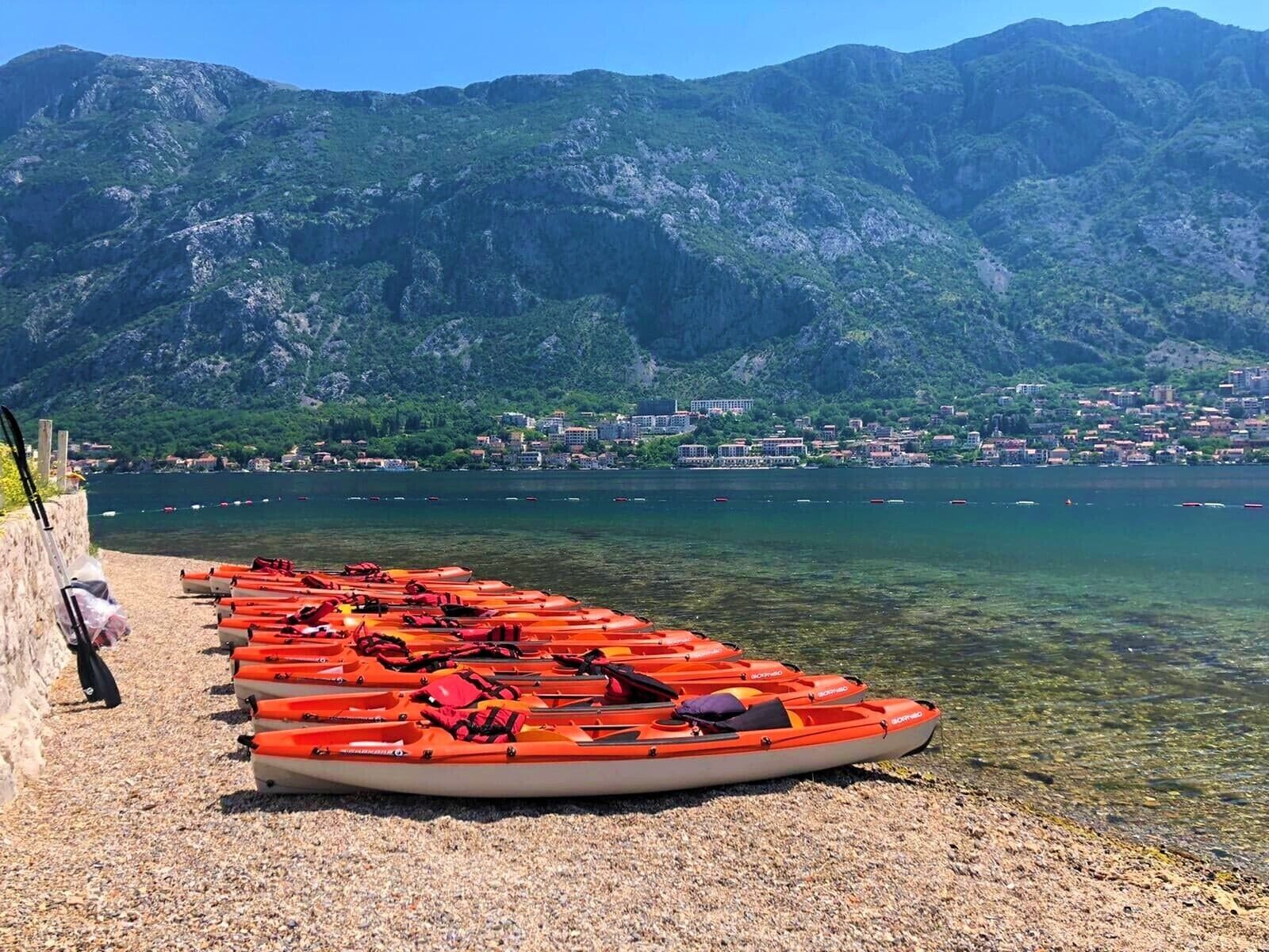 A row of orange kayaks lined up on a pebbled shore with a mountainous backdrop and a town across the water.