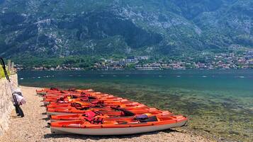A row of orange kayaks lined up on a pebbled shore with a mountainous backdrop and a town across the water.