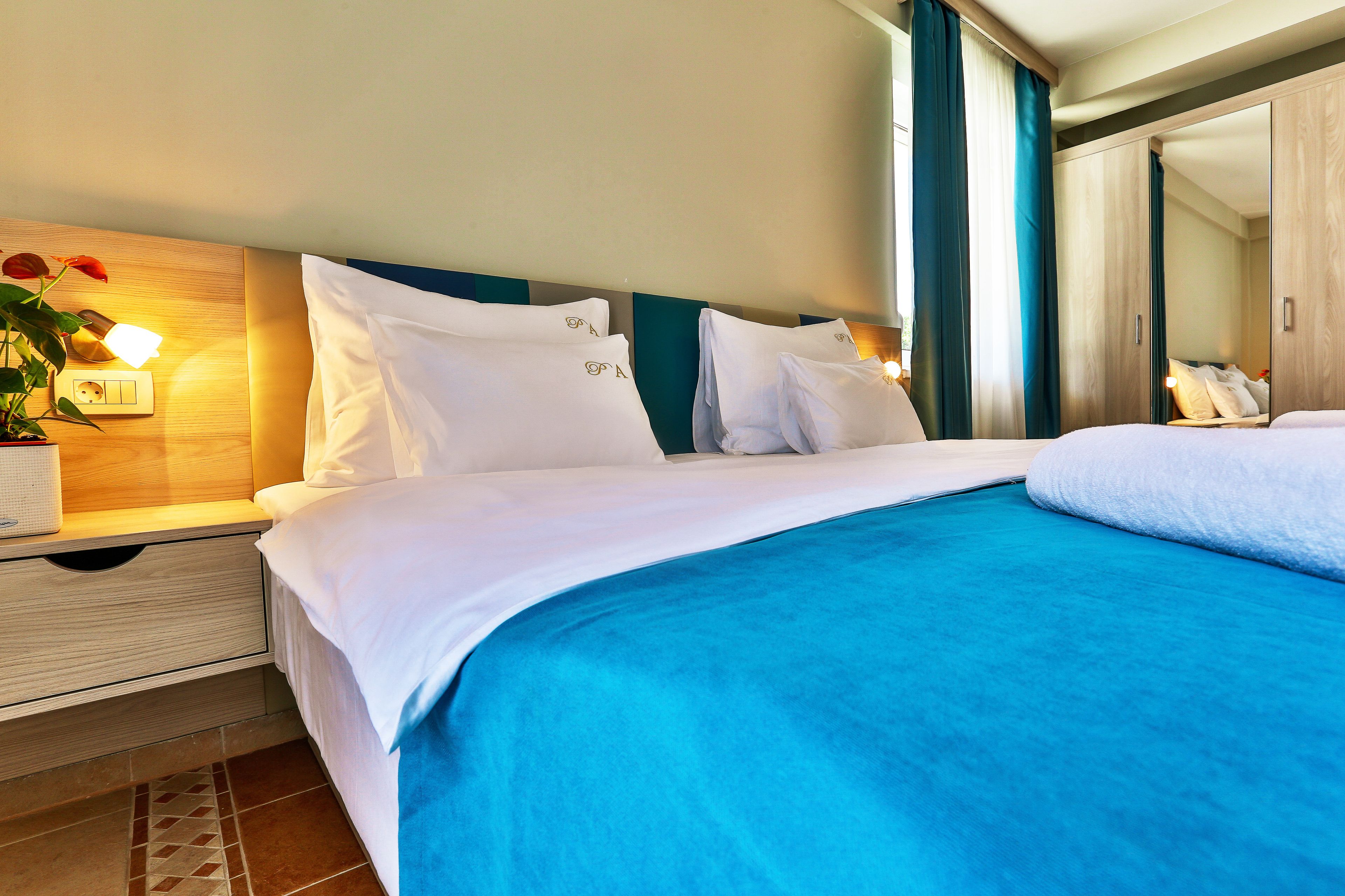 A hotel room with a bed, a nightstand with a lamp, a mirror, and a potted plant.