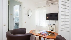 Ein Zimmer mit braunem Ledersessel, einem Glastisch mit einer Zeitschrift, einem Flachbildfernseher und einer kleinen, weißen Kommode mit einem Radio.