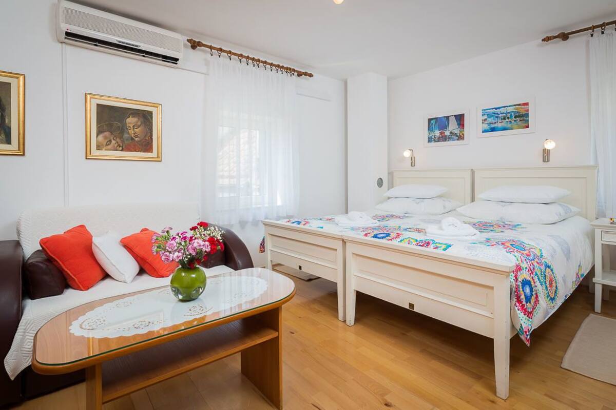 A bedroom with a double bed, a bedside table, a small table with a vase, a sofa with cushions, and a wall-mounted air conditioner.
