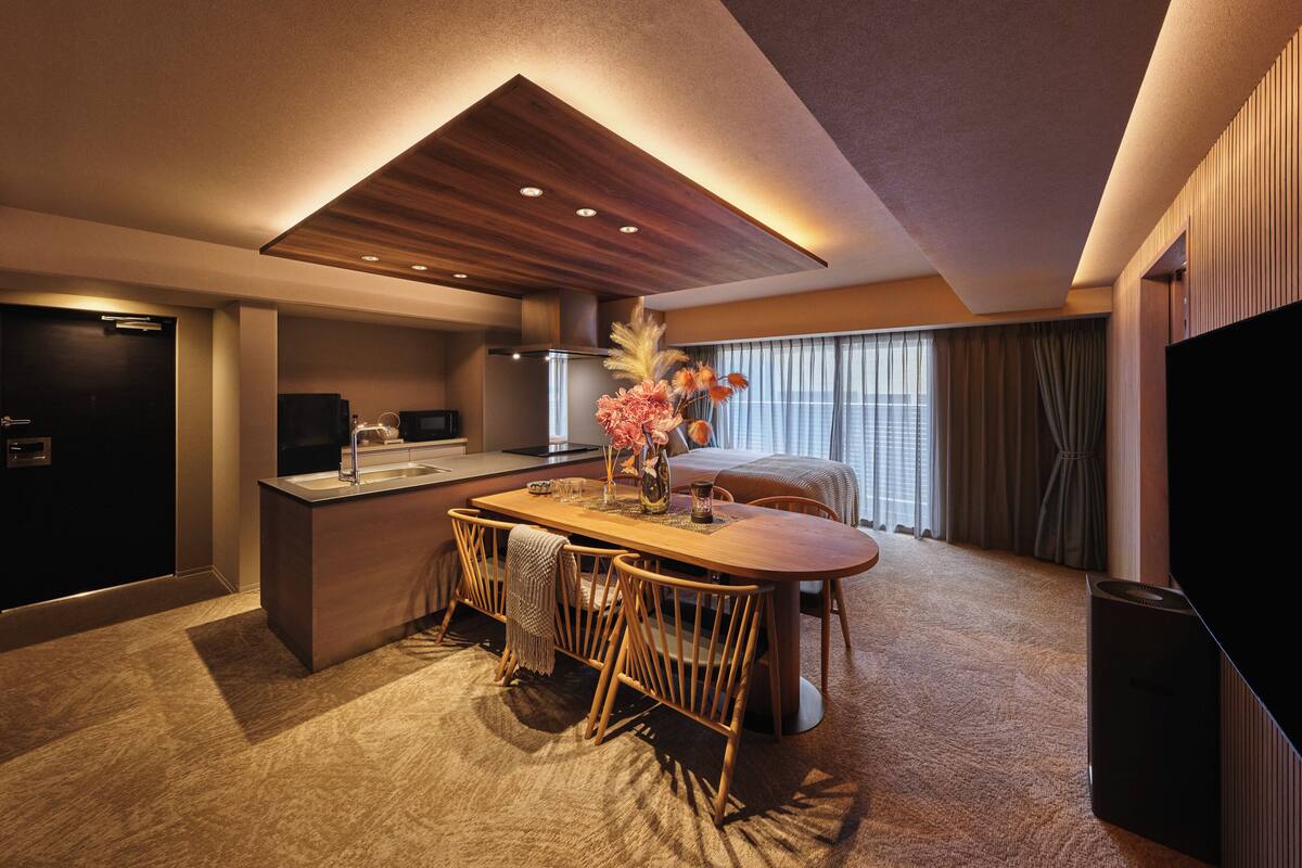 Une chambre d’hôtel moderne avec une table à manger en bois, un coin cuisine, un grand lit et un téléviseur.