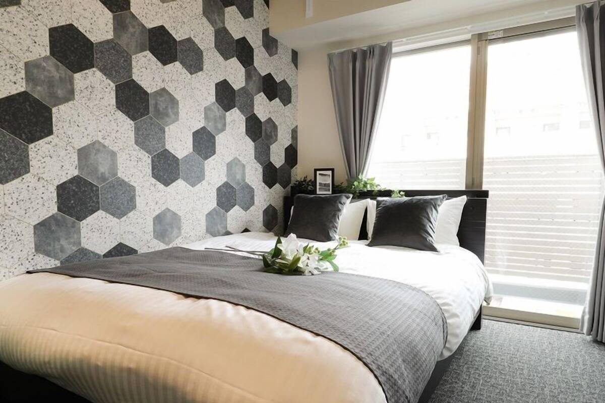 Une chambre d’hôtel moderne avec un mur orné d’un motif géométrique, un lit soigneusement fait avec une palette de couleurs en nuances de gris et de blanc, et une vue sur la ville à travers de grandes fenêtres.
