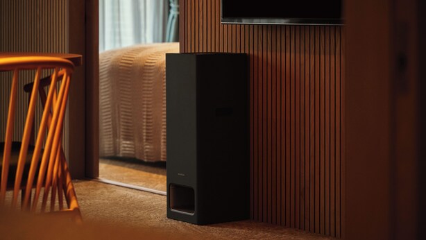 Een kamer met een bed, een houten stoel, een zwarte speaker en een aan de muur bevestigde televisie.
