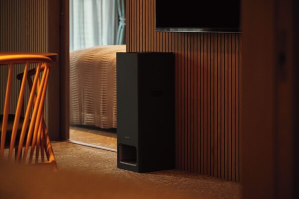 Une pièce comprenant un lit, une chaise en bois, un haut-parleur noir et un téléviseur fixé au mur.
